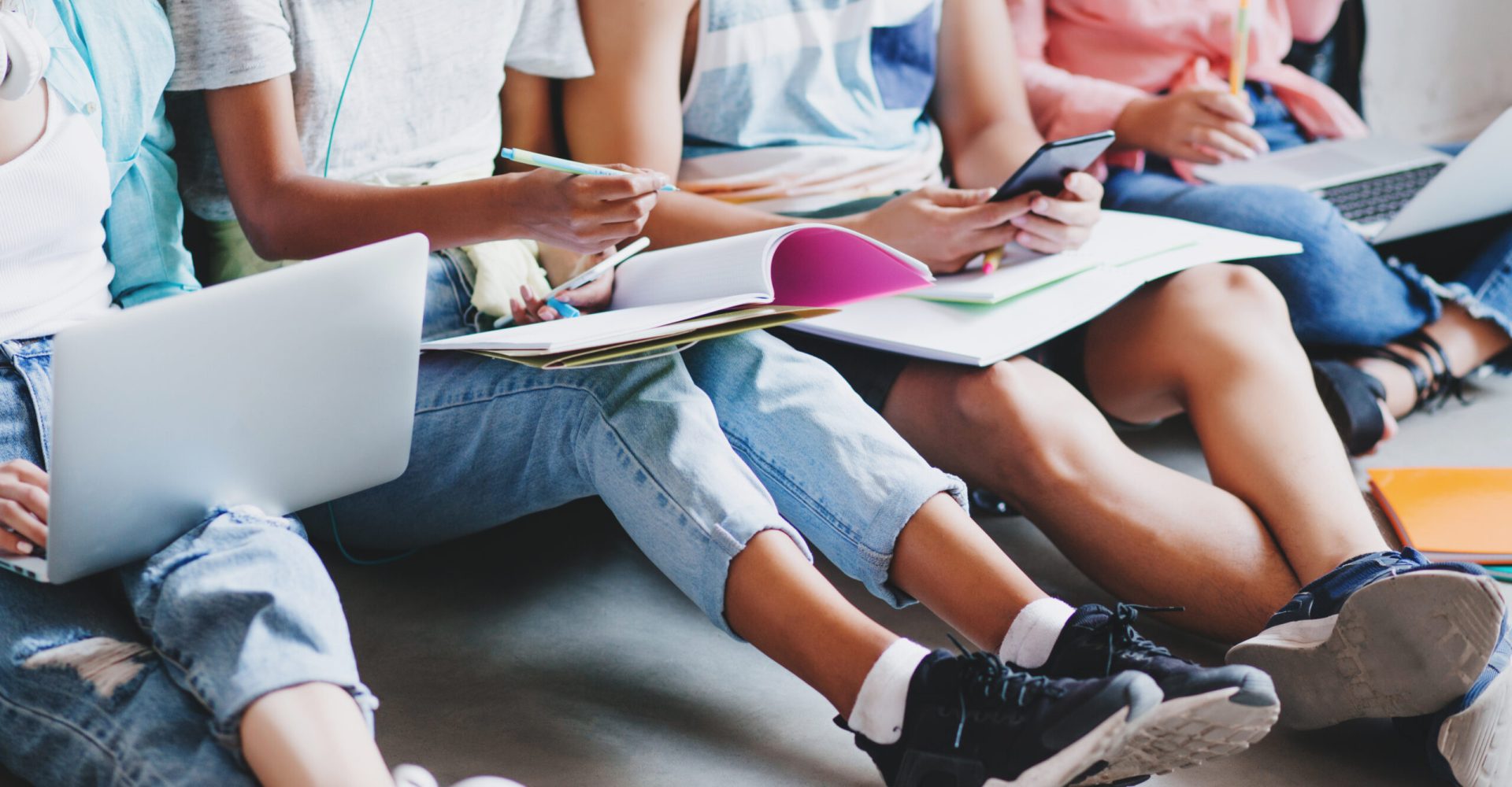 Girl in blue jeans and black sneakers writing lecture in big textbook, sitting on the floor with college friends. Young man typing message on phone while other students working with laptops..